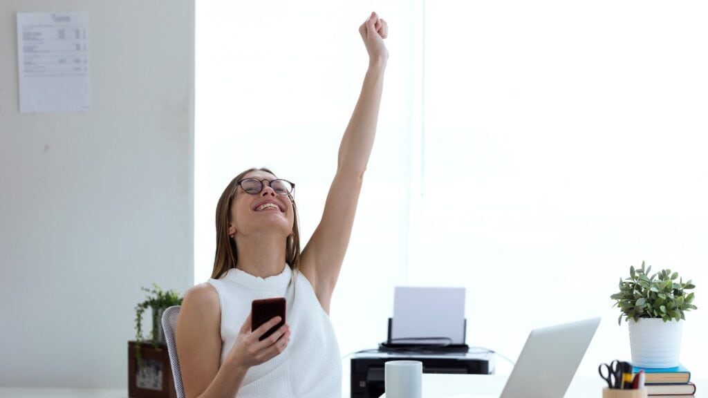 Joyful woman celebrating success with smartphone in modern office setting, representing social media engagement and digital marketing achievements.