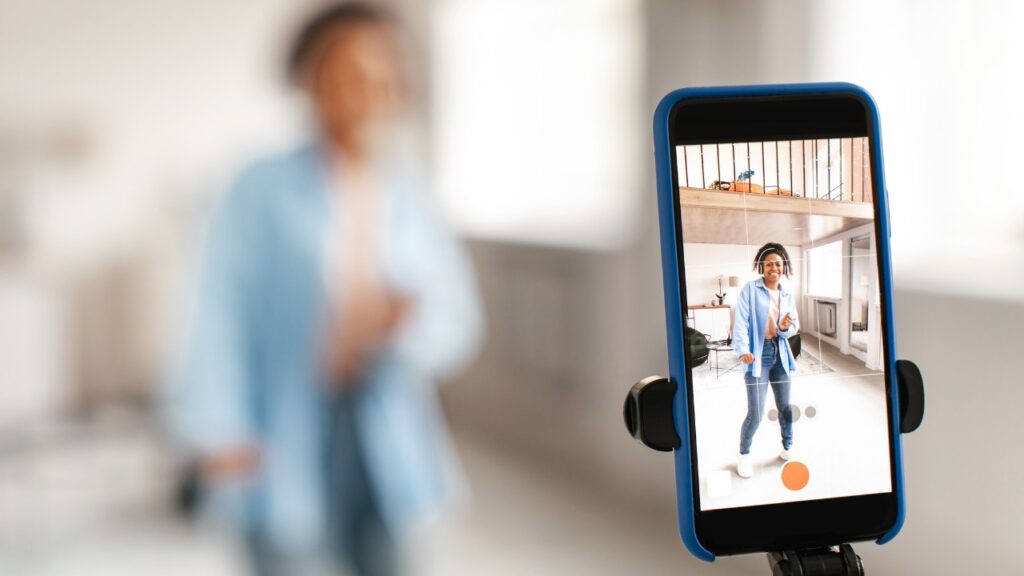 Vibrant young woman taking a selfie indoors with smartphone on tripod, smiling and enjoying social media content creation, showcasing modern lifestyle, creativity, and digital photo sharing.