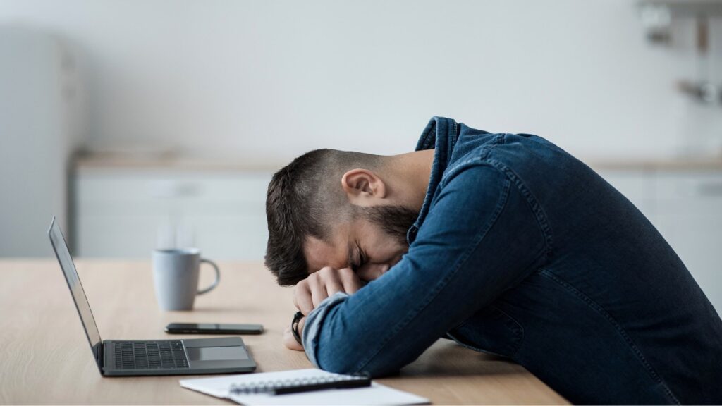Frustrated man overwhelmed with social media stress, sitting at desk, digital devices, workplace burnout, social media marketing difficulties, online marketing frustration, social media management challenges, internet marketing problems, social media strategy issues, digital marketing exhaustion.